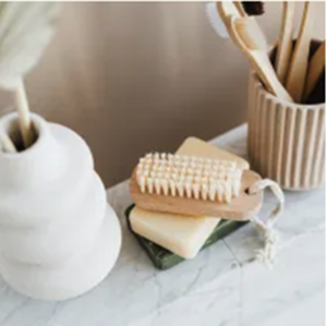 Close-up de produtos de banho e higiene sobre uma bancada de mármore, destacando uma escova de corpo de madeira sobre sabonetes em barra empilhados, um vaso branco com folhas secas e escovas de dente de bambu em um copo canelado.