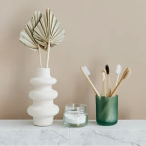 Arranjo minimalista de produtos de higiene pessoal em uma bancada de mármore, incluindo um vaso branco com folhas secas, um pequeno pote de vidro com pó branco (possivelmente dental) e escovas de dente de bambu em um porta-escovas de vidro verde.
