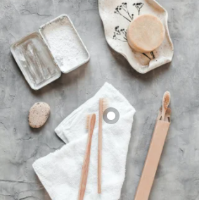 Vista aérea de produtos de higiene pessoal sustentáveis sobre uma superfície cinza texturizada, incluindo escovas de dente de bambu, um sabonete sólido, um sabonete em pedra, uma toalha branca e uma lata aberta com pó.