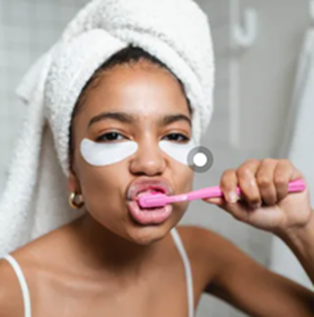 Mulher jovem com uma toalha na cabeça e adesivos de tratamento sob os olhos (eye patches) escovando os dentes com uma escova de dente rosa.