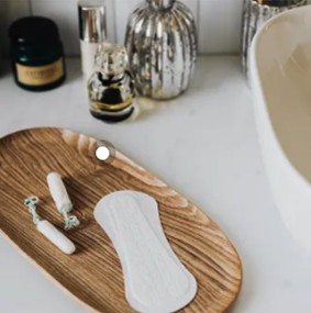 Produtos de higiene feminina, incluindo absorventes e tampões, dispostos em uma bandeja de madeira perto de um lavatório e frascos de produtos de beleza em segundo plano.