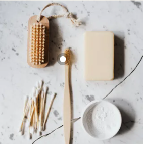 Disposição de produtos de higiene e cuidados pessoais em uma bancada de mármore branco. Itens incluem uma escova de corpo de madeira, uma barra de sabonete, cotonetes, uma escova de dente de bambu e uma tigela com pó branco.